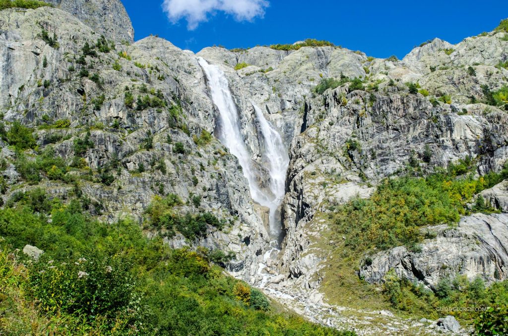 Cascade jaillissant dans un ravin boisé, montagnes en arrière-plan et sentier de trekking visible