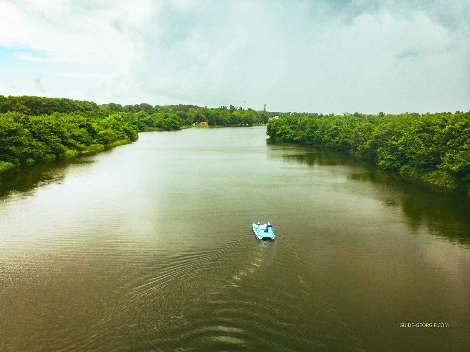 Deux personnes assises dans un pédalo bleu sur une étendue d'eau, ciel nuageux, végétation en arrière-plan