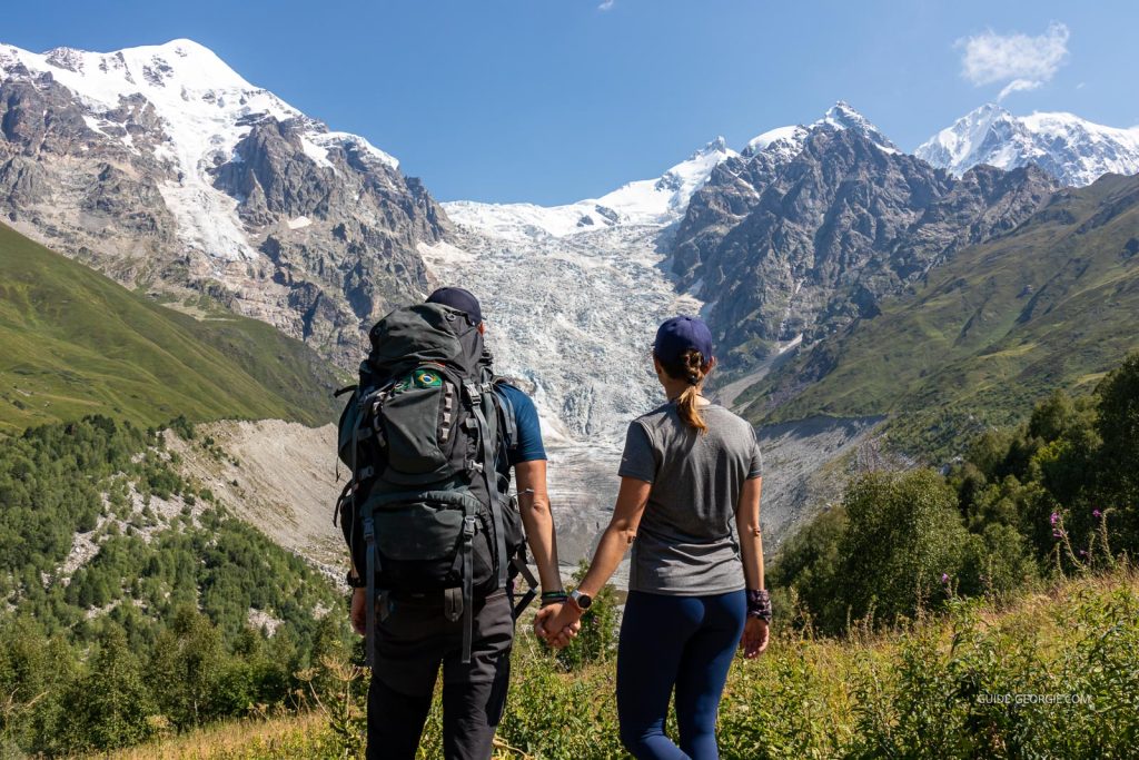 Un couple debout sur un sentier regarde des sommets enneigés et un glacier à l’horizon dans le massif du Grand Caucase