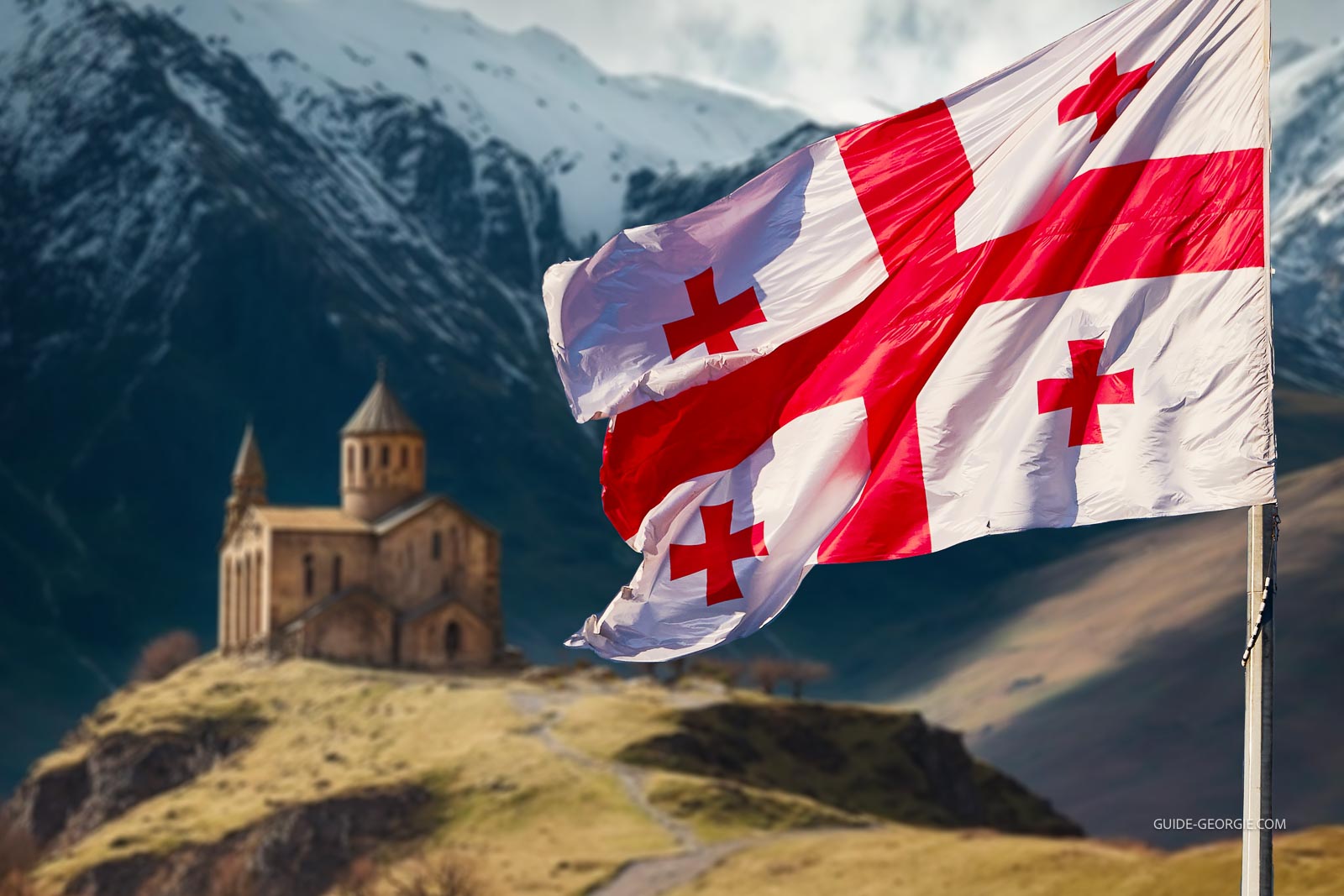 Drapeau national de la Géorgie flottant devant des montagnes enneigées et une église en architecture géorgienne traditionnelle