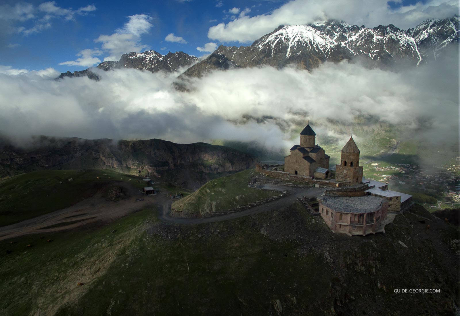 Vue aérienne de l'église de la Trinité à Gergeti au premier plan, montagnes enneigées et nuages en arrière-plan