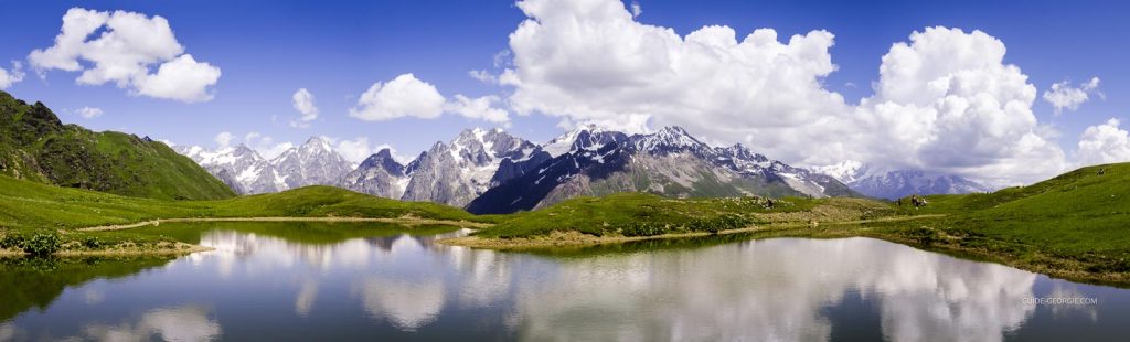 Lac de montagne aux eaux calmes entouré de sommets herbeux et de rochers, ciel nuageux au-dessus