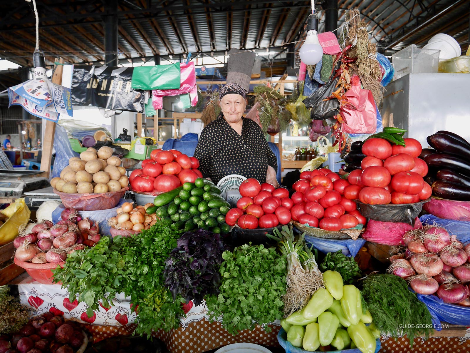 Étals de marché en plein air avec divers légumes colorés (concombres, aubergines, tomates) disposés sur des caisses et serviettes