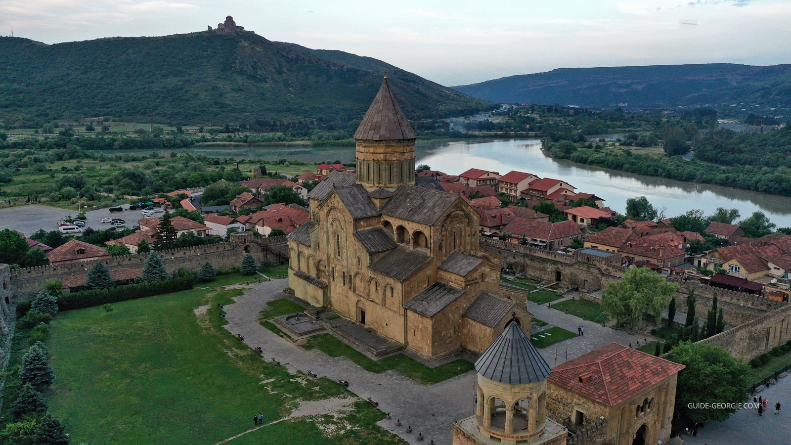 Vue extérieure du monastère Svetitskhoveli, bâtiment religieux en pierre avec coupole(s) et toits, ciel bleu et végétation alentour