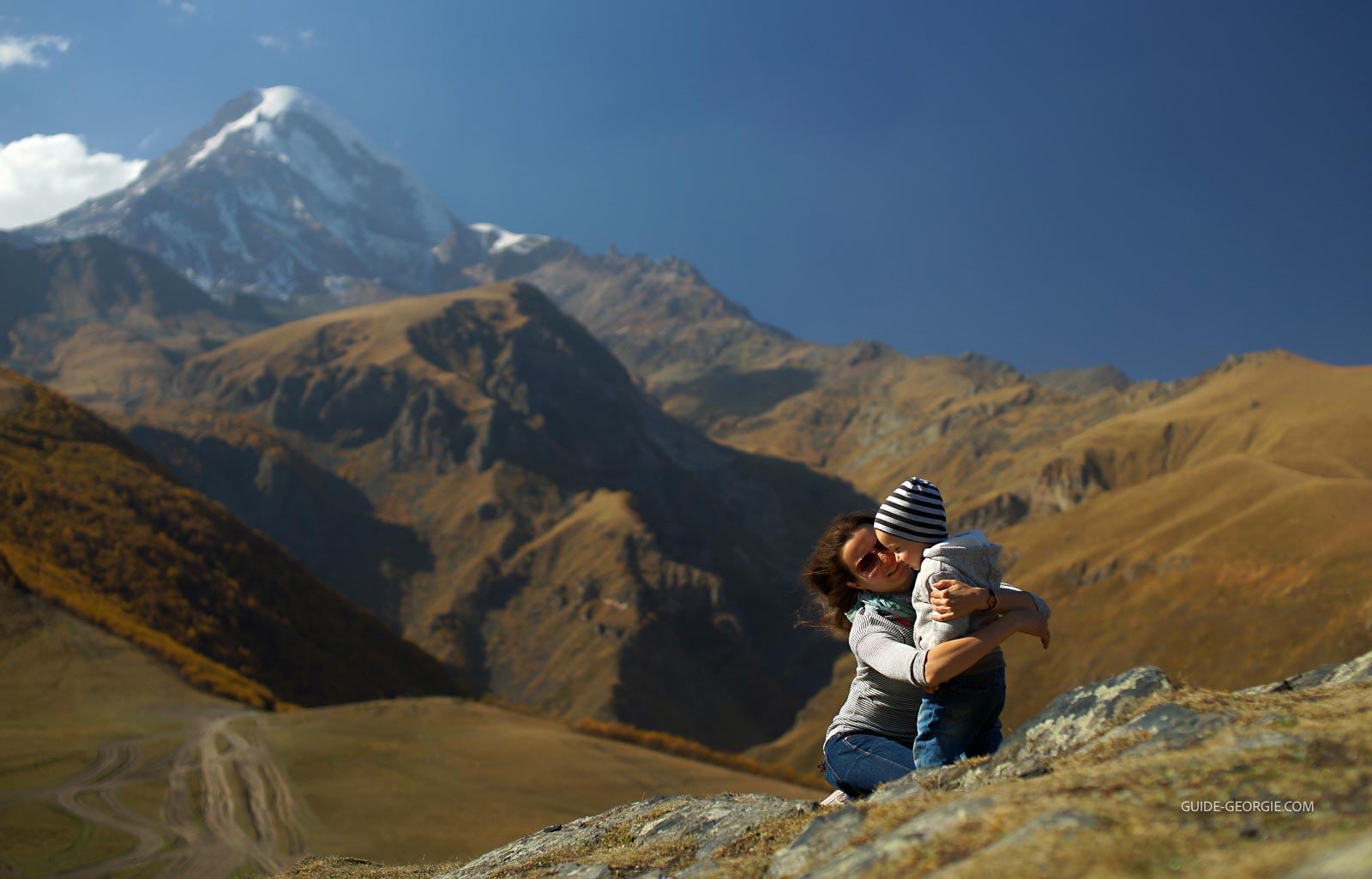Vue arrière d'un petit garçon et de son père regardant un large glacier enneigé, collines brunes en arrière-plan et ciel bleu