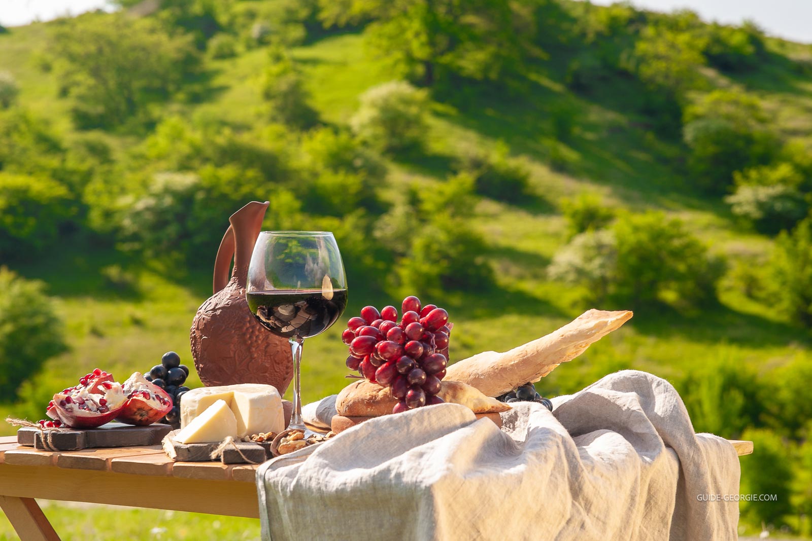 Table de pique-nique en bois dans une prairie, avec bouteille de vin rouge, fromages, fruits, raisins et pain sur la table