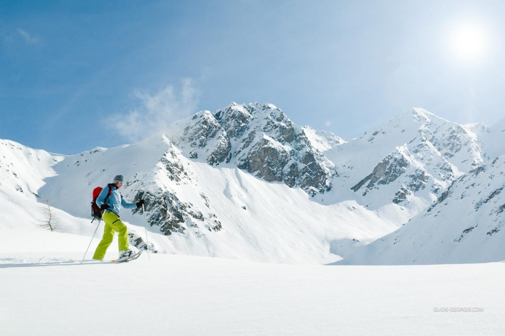 Femme en tenue d'hiver marchant en raquettes sur un sentier enneigé en montagne, bâtons de marche visibles, ciel et nuages en arrière-plan