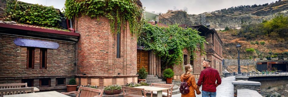 Femme en veste brune et homme en chemise rouge marchant dans une rue ancienne bordée de bâtiments historiques à Tbilissi