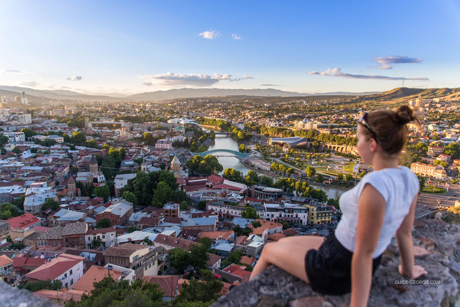 Vue aérienne d'un quartier ancien de Tbilissi avec une touriste souriante, bras croisés, et toits de la ville en arrière-plan